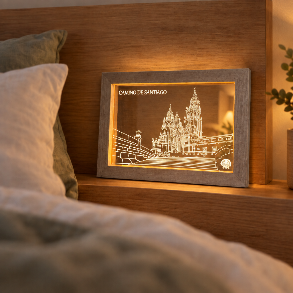Grey-washed Camino de Santiago LED frame standing lit on a wooden bed headboard shelf in a cozy bedroom, the engraved art glowing on a clear acrylic panel.