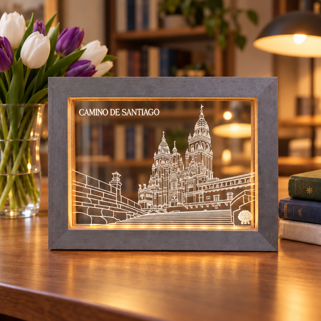 Grey-washed Camino de Santiago LED frame lit on a wooden desk in a cozy study with white and purple tulips, books, and a warm lamp.