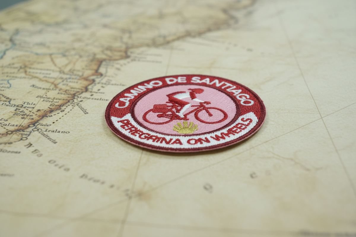 Red and pink Peregrina On Wheels iron-on patch from Pilgrim Artifact lying flat on an antique map of the Camino routes 