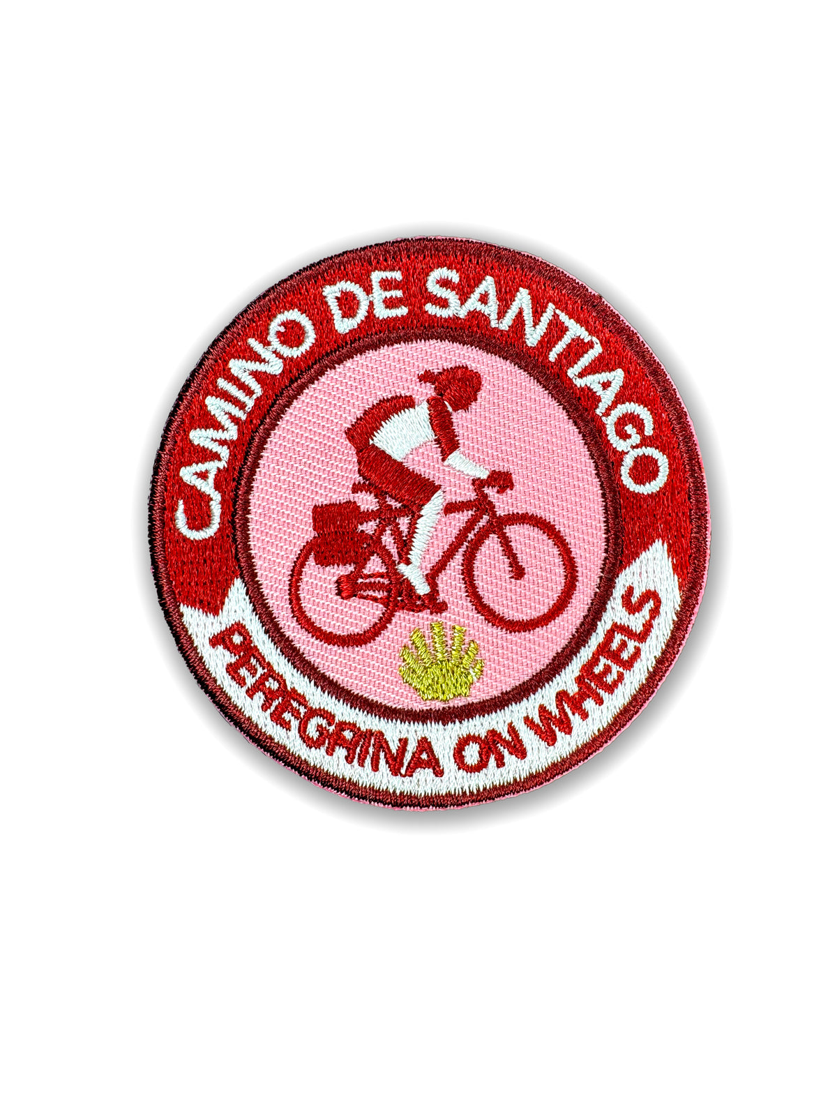 Pilgrim Artifact Peregrina On Wheels embroidered iron-on patch featuring a woman cyclist on a touring bicycle against a pink center, a gold scallop shell, and a red and white Camino de Santiago border, shown on a white background. 